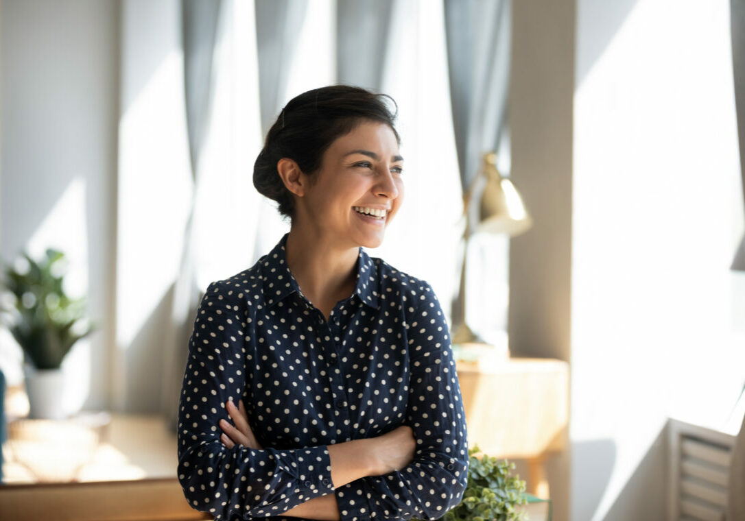 Smiling young Indian woman stand look in distance window dreaming or visualizing, happy overjoyed millennial ethnic girl thinking or planning of future success opportunities at home